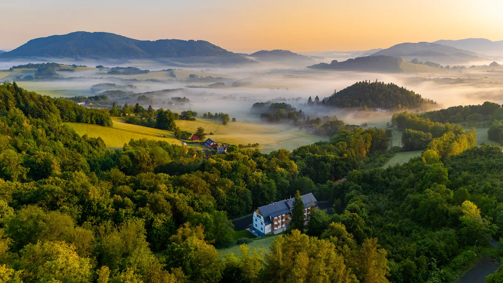Hotel Edvard z výšky za východu slunce - bílá budova s balkony v lesích na Horečkách, v údolí mlha, v pozadí hřebeny Beskyd