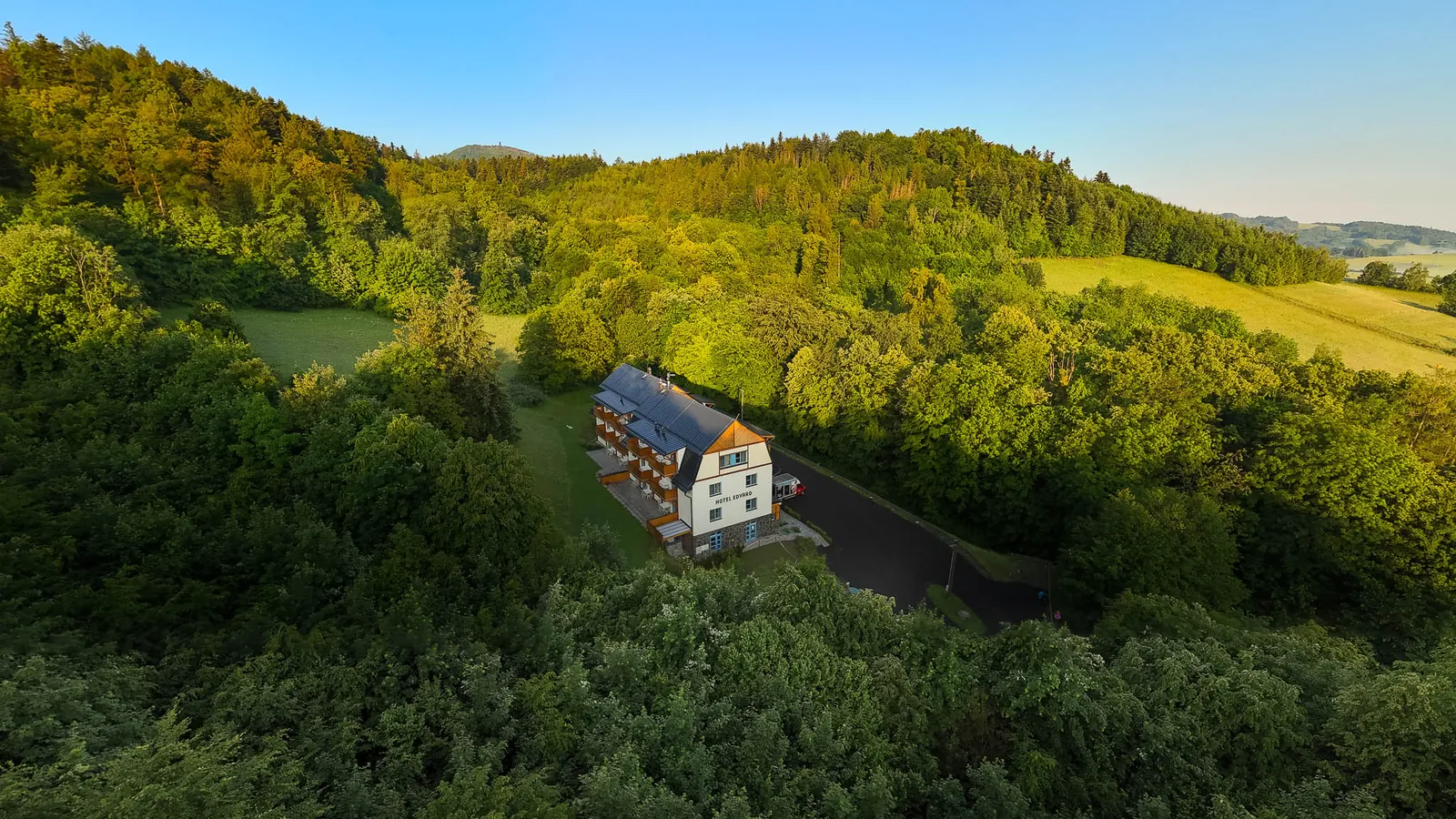 Hotel Edvard v létě z výšky - bílá budova s balkony a příjezdovou cestou uprostřed zeleného lesa, za ní louky a kopce pod modrým nebem