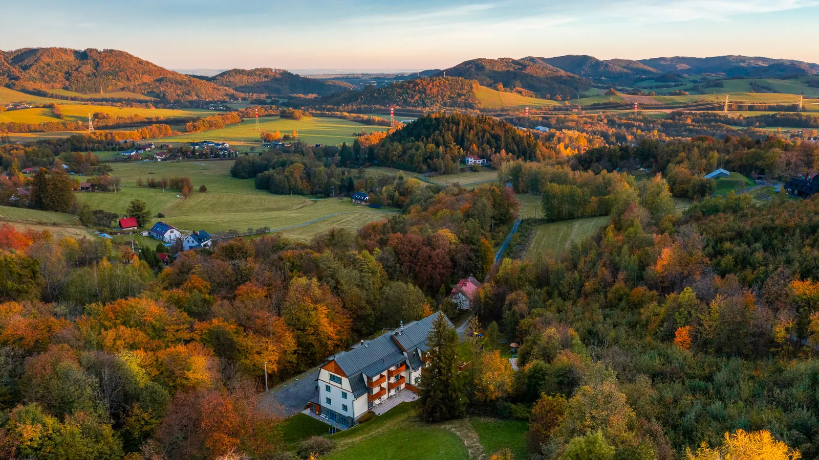 Hotel Edvard na podzim z výšky - bílá budova s dřevěnými balkony mezi barevnými stromy, za ní louky a hřebeny Beskyd ve večerním světle